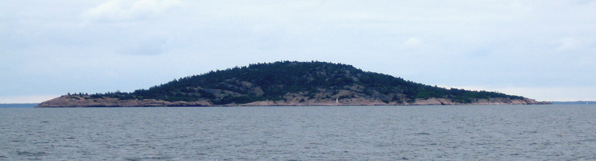 The dome-shaped granite island of Blå Jungfrun seen from the sea, with a rocky shoreline and dense forest on top.