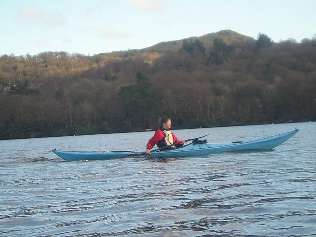 A person kayaking on calm water wearing a visible life vest, with wooded hills in the background.