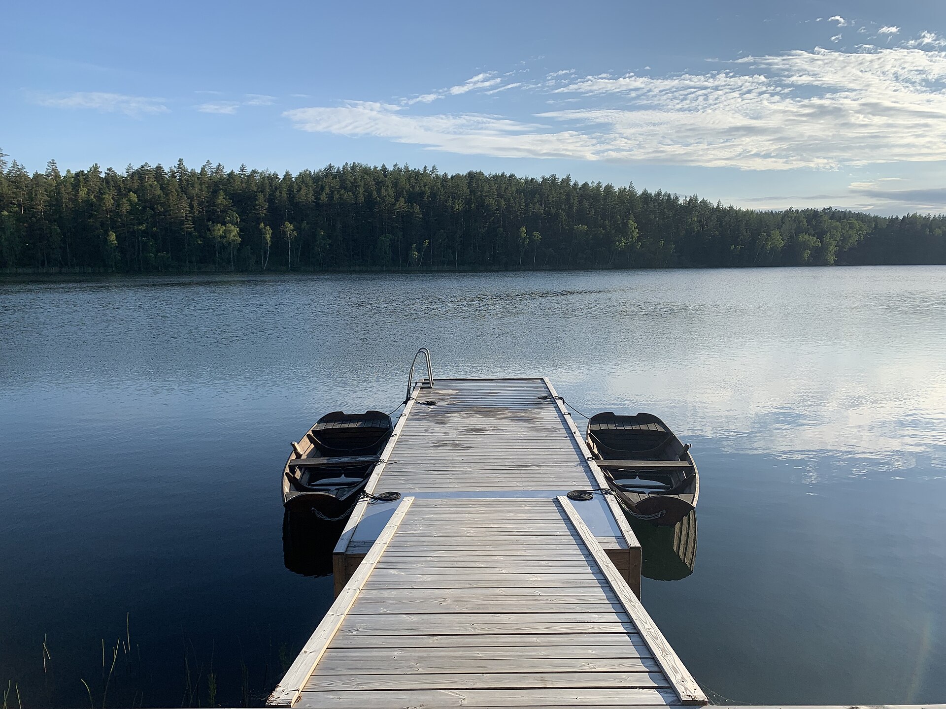 Träbrygga med två roddbåtar vid en lugn sjö norr om Västervik, med skogklädd strand i bakgrunden.